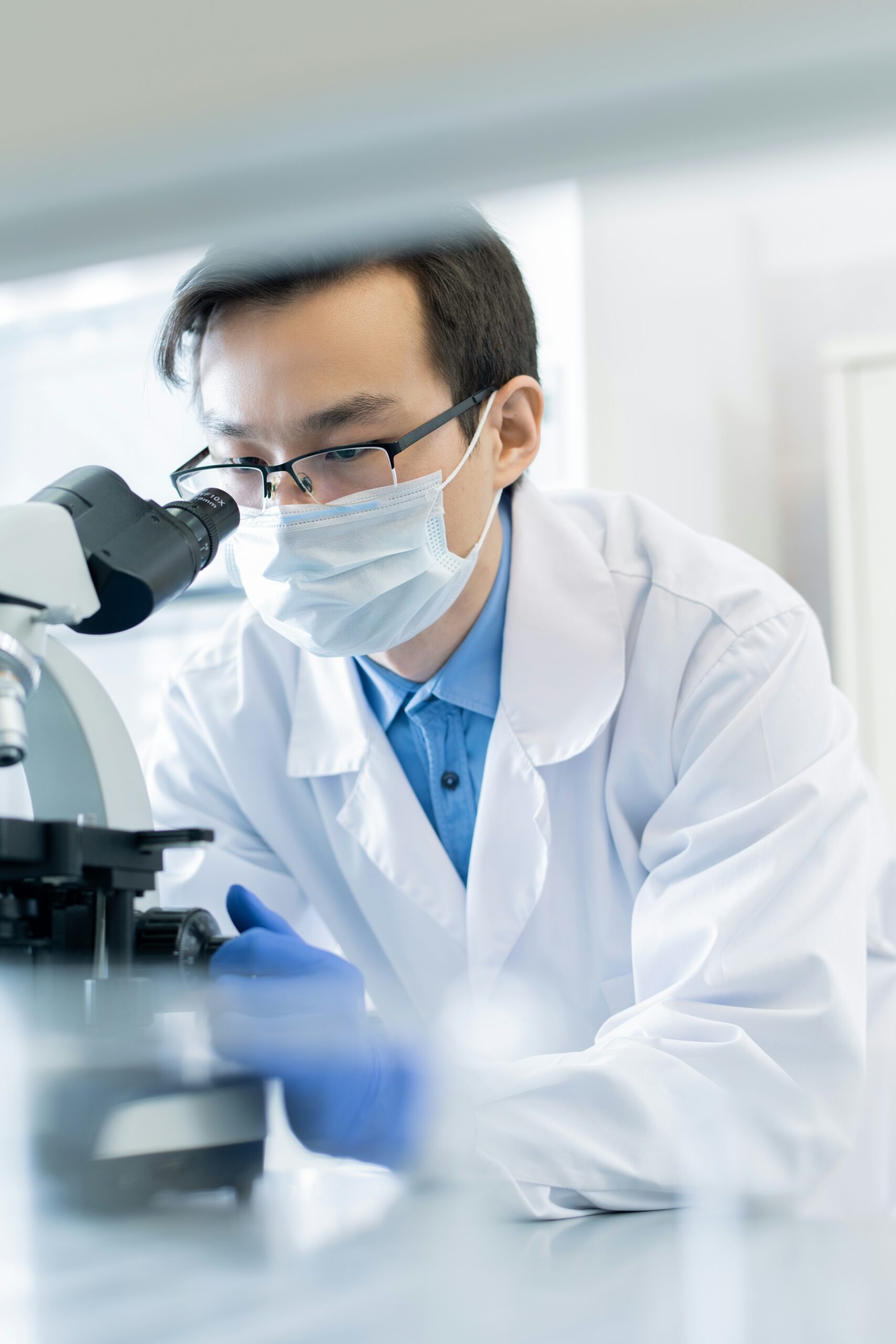 Vertical portrait of young Asian pharmacologist wearing protective mask examining specimen chemical reaction using microscope in modern laboratory